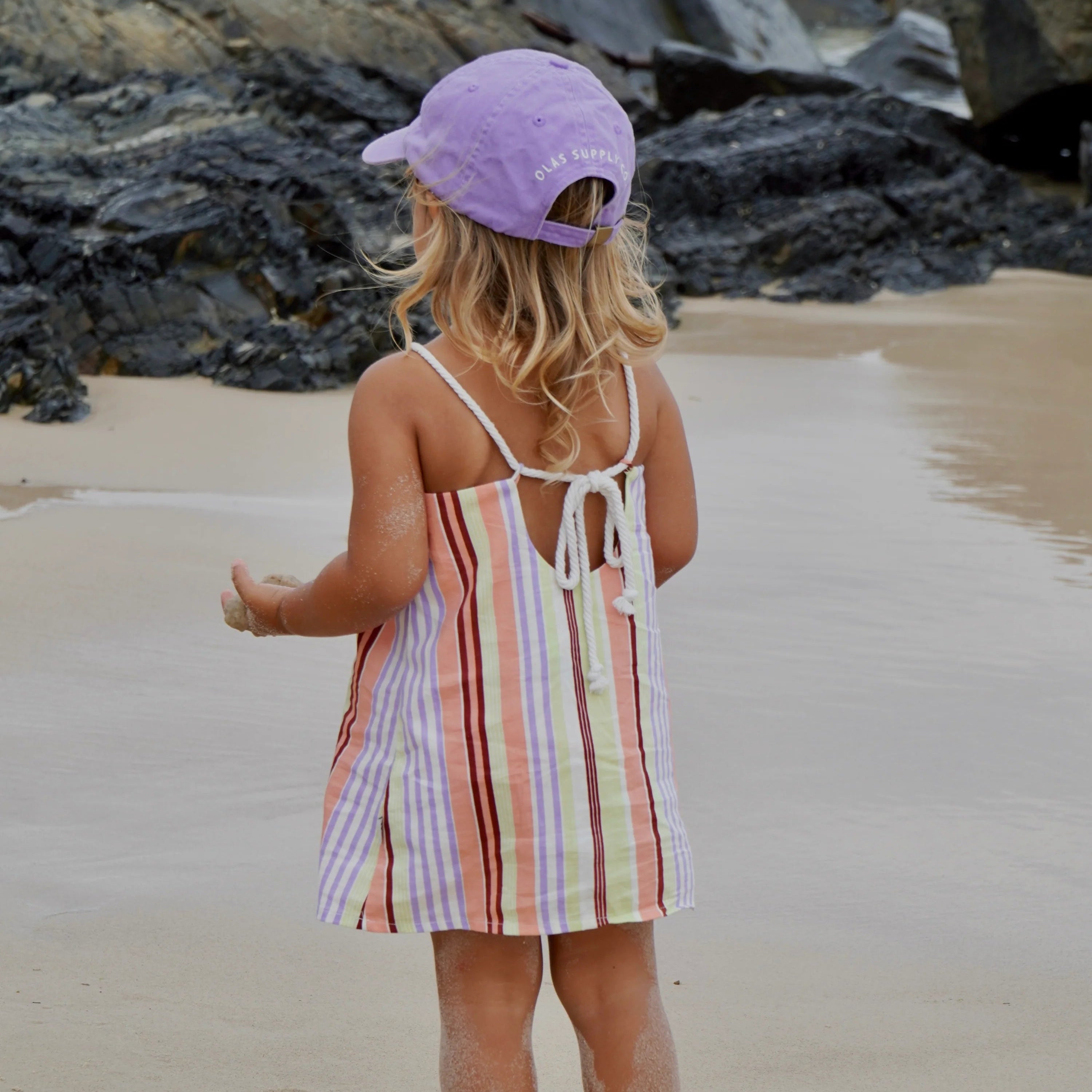 Child in a striped dress and purple cap standing on a beach with rocks and water in the background